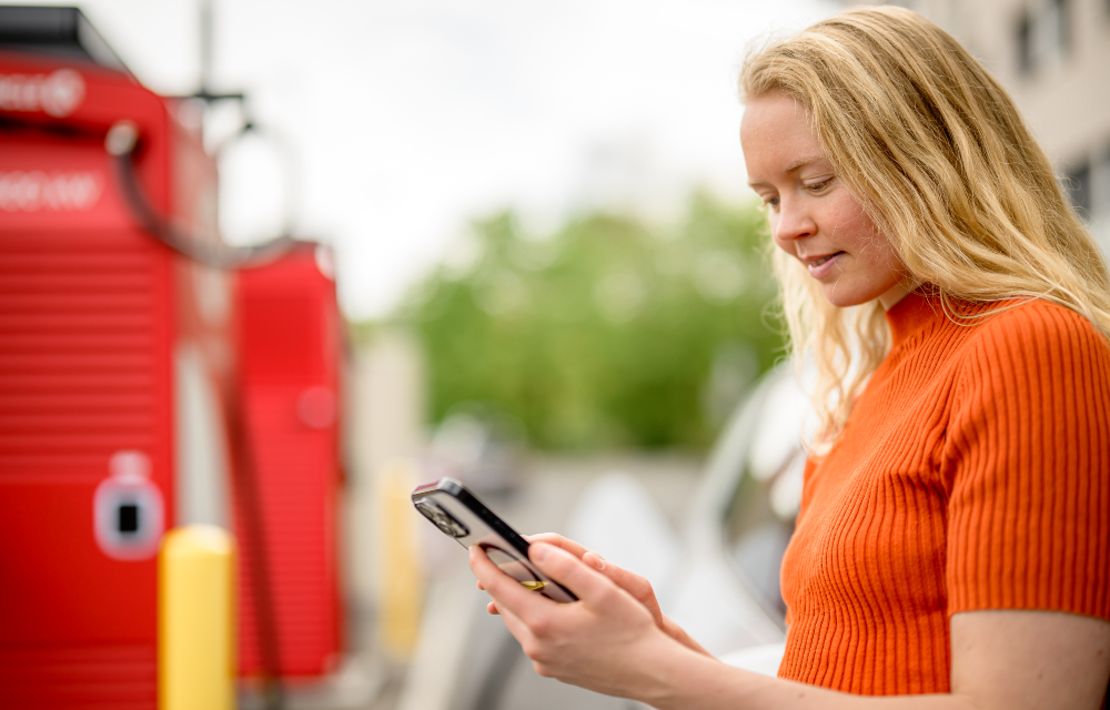 Kvinde i orange trøje står udendørs ved en ladestation og ser på sin mobiltelefon