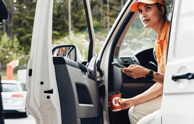 Woman getting out of her van at a Circle K station, with her phone in one hand and a coffee in the other.